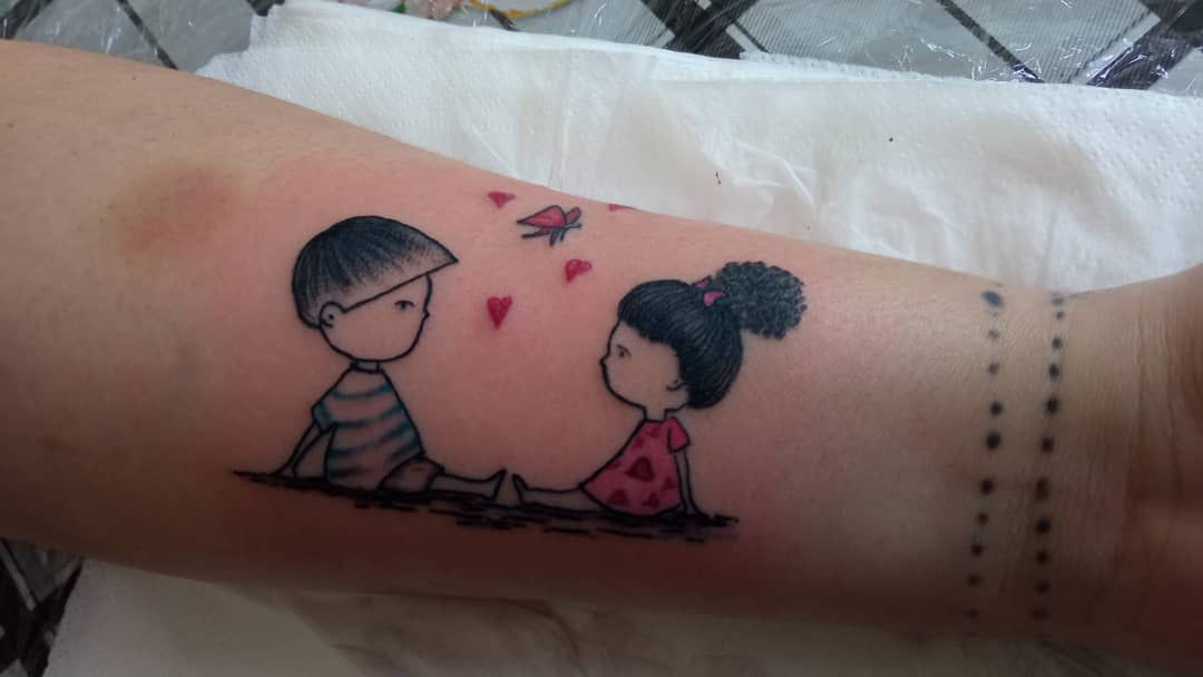 Tattoo of a boy and girl sitting with two hearts and a butterfly above on an arm